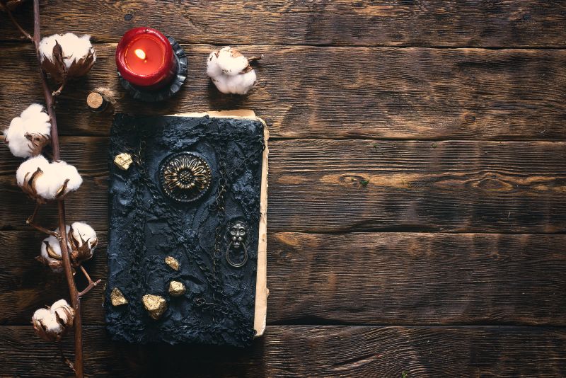 Stara magiczna księga leżąca na drewnianym stole. Obok niej zapalona świeca i kulki bawełny.